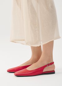 Des chaussures slingback en cuir rouges avec un bout pointu, une semelle plate et une sangle de cheville réglable, associées à un vêtement transparent beige.