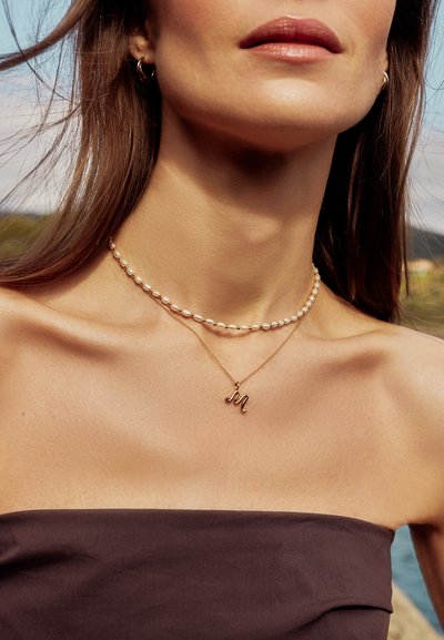 Γυναίκα που φοράει κολιέ με μαργαριτάρια και χρυσό κολιέ με μενταγιόν το γράμμα "μ", ντυμένη με στράπλες σκούρο καφέ τοπ, έξω δίπλα σε νερό.