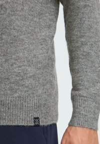 Suéter de lana gris con dobladillo y puños acanalados, y con una pequeña etiqueta negra en la parte inferior. La textura parece suave y ligeramente difusa.