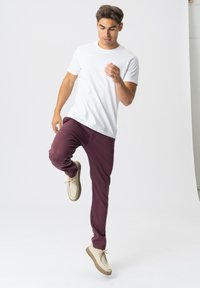 Hvid kortærmet t-shirt parret med bordeaux joggerbukser og lyse sko. Modellen er i en dynamisk positur og løfter det ene ben.