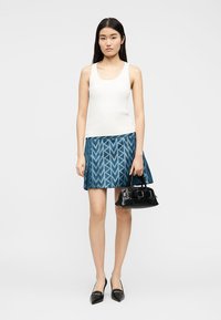 Jeune femme aux cheveux longs, portant un haut blanc sans manches et une jupe bleue à motifs, tenant un sac à main noir, debout devant un fond uni.