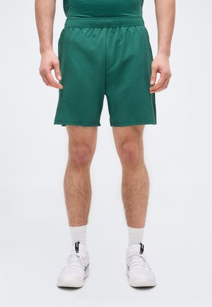 Férfi sötétzöld sportnadrágban, fehér térdzokniban és fehér futócipőben, sima világos háttér előtt állva.