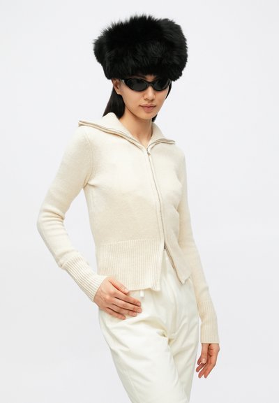 Kremfarget strikket cardigan med glidelås, ribbestrikket nederkant og krage. Brukt med en fluffy svart hatt og mørke solbriller. Kombinert med lyse bukser.