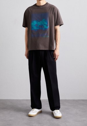 Grijze katoenen T-shirt met een grafisch ontwerp van een blauwe berg, ontspannen pasvorm, gecombineerd met zwarte geplisseerde broeken en witte sneakers met beige zolen.