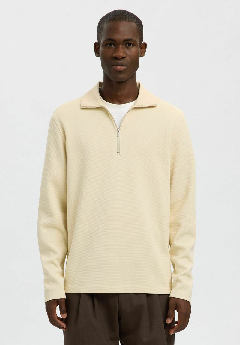 Beige svetr s dlouhým rukávem a límcem na zip, vyrobený z texturované látky. Nositý přes bílé tričko ve spojení s hnědými kalhotami.
