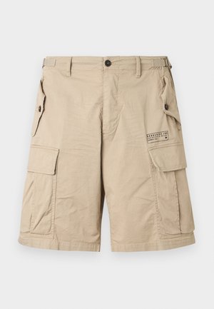 Bēša krāsas kravas īsās bikses ar vairākiem kabatām, kurām ir pogas, regulējamas sānu lences, jostu cilpas un mazu drukātu logotipu labās augšstilba kabatas vietā.