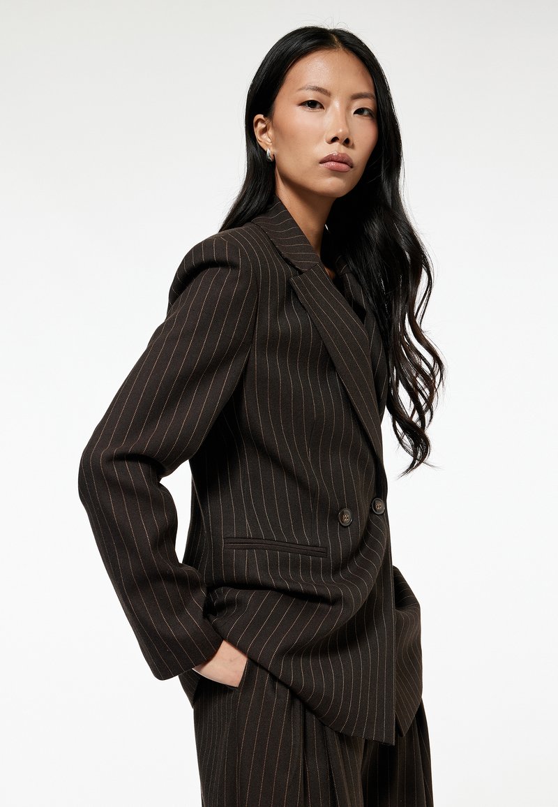 Blazer de corte cruzado en marrón oscuro con rayas, con hombreras y bolsillos frontales, que presenta líneas naranjas en contraste para un detalle adicional.