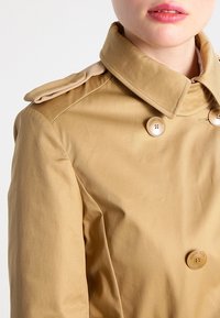 Trench coat beige con design doppiopetto, grandi bottoni, spalline e una texture liscia. Vestibilità sartoriale con colletto strutturato.