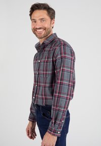 Grijze geruite shirt met rode en blauwe strepen, met knoopsluiting kraag, lange mouwen en twee borstzakken, gedragen met donkerblauwe jeans.