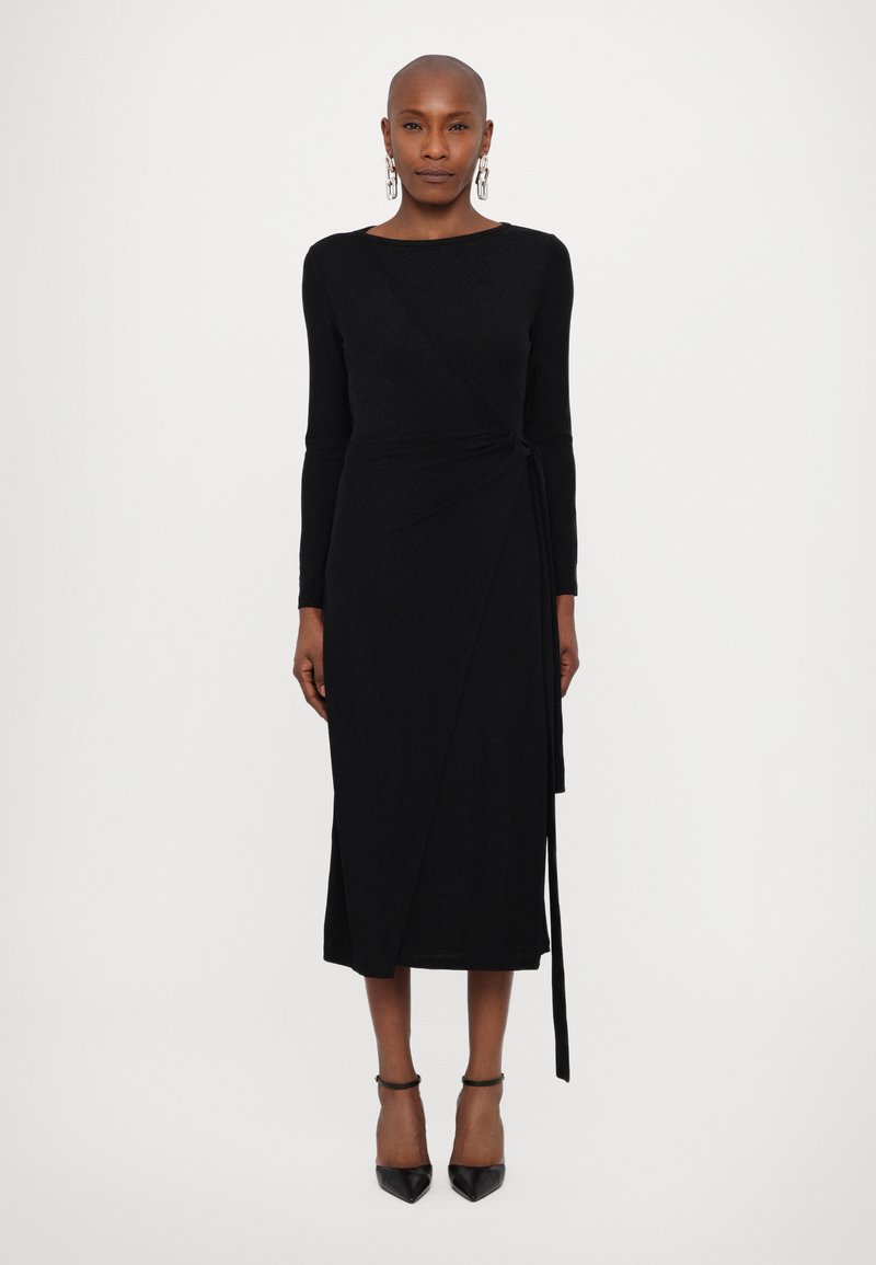 Vestido envolvente negro de manga larga con un diseño ajustado, dobladillo asimétrico y detalle de lazo lateral. Textura suave y longitud midi, combinado con tacones de punta.