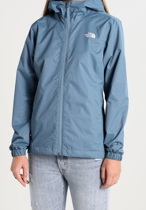 Kobieta ubrana w niebieską kurtkę The North Face zapinaną na zamek z elastycznymi mankietami, w połączeniu z jasnoszarymi, postarzanymi jeansami, stojąca na jednolitym tle.