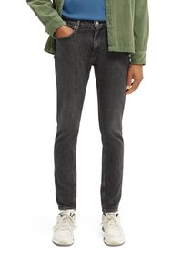 Jeans de mezclilla negra con corte ajustado, que presentan una textura desgastada y un diseño de cinco bolsillos, complementados con una camisa azul y una chaqueta verde.