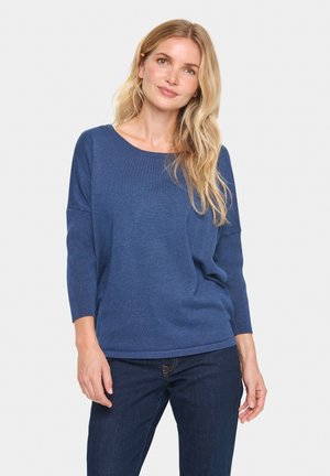 Woman with long blonde hair wearing a blue long-sleeve sweater and dark blue jeans, standing against a plain light background.