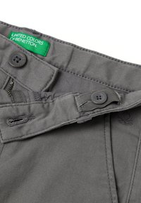 Pantalones grises con cremallera, cierre de botón y etiqueta verde "United Colors of Benetton". Presentan costuras acentuadas y una textura suave.