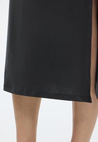 Μαύρο φόρεμα μάκρος μέχρι το γόνατο από απαλό ύφασμα, με πλαϊνή σχισμή και ευθεία γραμμή στο τελείωμα. Μινιμαλιστικός σχεδιασμός χωρίς εμφανή σχέδια ή διακοσμήσεις.