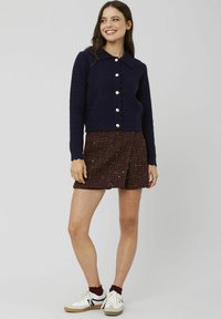 Cardigan en tricot bleu marine avec un ourlet festonné et de gros boutons, associé à une jupe à motifs bordeaux et des baskets blanches avec des accents noirs.