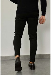 Pantalons noirs fuselés en tissu extensible, présentant une texture lisse et une silhouette étroite, associés à des chaussures en cuir foncé.