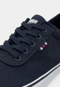 Mornarsko platneno tenisko z teksturirano površino, okrogli vezalni trakovi, črne odprtine za vezalke in majhna večbarvna oznaka. Bele gumijaste podplate naglašajo.