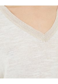 Kevyt, kermaisenvalkoinen paita, jossa on tekstuuri ja V-pääntie. Pääntiellä on kontrastivärinen, kiiltävä beige reunus.