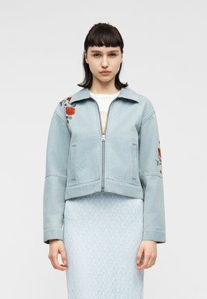 Junge Frau mit kurzen dunklen Haaren, die eine hellblaue Jeansjacke mit roter Rosenstickerei und einen hellblauen Spitzenrock trägt und vor einem schlichten Hintergrund steht.