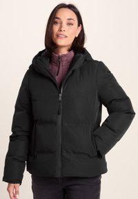 Woman wearing a black hooded puffer jacket over a purple zip-up sweater, standing with one hand in jacket pocket against beige background.