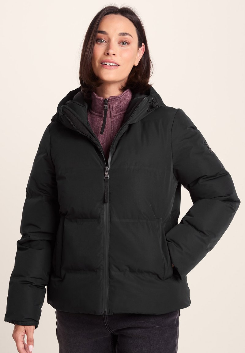 Woman wearing a black hooded puffer jacket over a purple zip-up sweater, standing with one hand in jacket pocket against beige background.