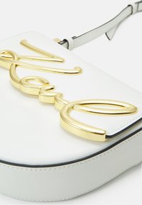 Biała torba na ramię z złotym metalowym logo. Gładka tekstura o zorganizowanym kształcie i kontrastowym czarnym wykończeniu. Regulowany pasek na ramię.