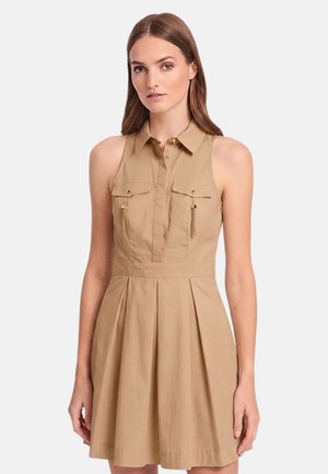 Donna che indossa un vestito camicia beige senza maniche con colletto, con bottoni sul davanti e due tasche sul petto con dettagli a nappine.
