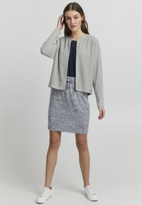 Ljusgrå texturerad cardigan med rund halsringning, bärs över en mörk topp och en melerad grå kjol, i kombination med vita sneakers.
