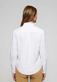 Camicia bianca a maniche lunghe con colletto sagomato, orlo curvo e polsini abbottonati; texture morbida con design classico.
