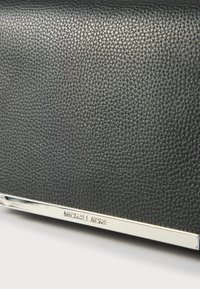 Melnas teksturētas ādas maciņš ar sudraba tonī metāla akcentu, kas priekšpusē ir iegravēts ar zīmola nosaukumu "MICHAEL KORS".