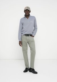 Szary pulower z suwakiem do połowy, zielone spodnie, czarne buty, beżowa czapka z daszkiem. Gładka tkanina o dopasowanym kroju, z logo na piersi.