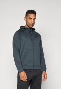 Mørkegrønn zip-up hettegenser laget av mykt, elastisk materiale. Har hette, kengurulommer og en liten logo på brystet.