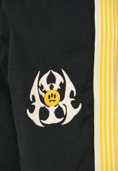 Μαύρο ύφασμα με έντονο κεντημένο σχέδιο σε λευκό, featuring αιχμηρά σχήματα και ένα κίτρινο χαμογελαστό πρόσωπο, δίπλα σε κίτρινες και λευκές ρίγες.