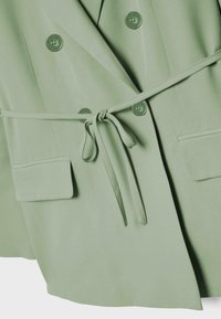 Light green double-breasted blazer with four buttons, front pockets with flaps, and a tied fabric belt at the waist.