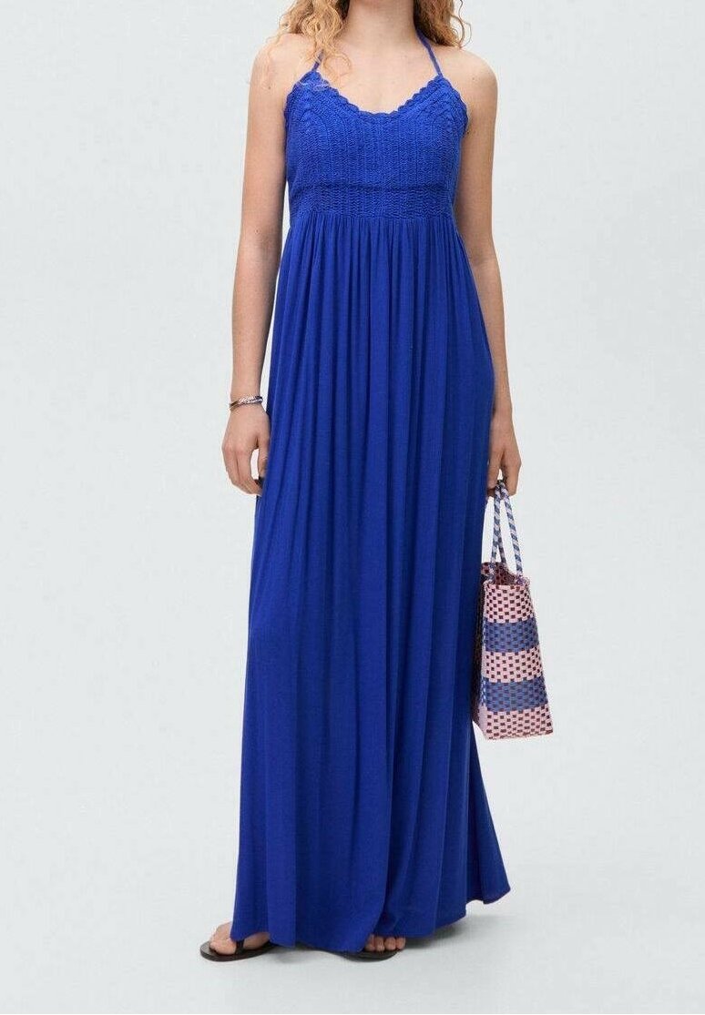 Femme portant une robe longue bleue sans manches, tenant un sac tissé rayé rose et bleu, debout devant un fond uni.