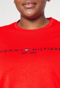 Rød bomulds-t-shirt med rund halsudskæring og et trykt logo i marineblå, der inkluderer "TOMMY HILFIGER EST. 1985".