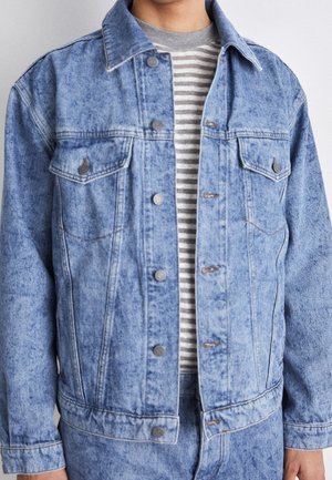 Mężczyzna w jasnoniebieskiej kurtce acid wash z denimu, noszący pod nią szarą i białą pasiastą koszulę, stojący na jednolitym tle.