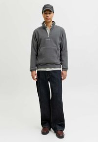 Pull en polaire gris avec zip moitié et poche avant, superposé sur une chemise à carreaux. Associé à un jean large sombre et des chaussures brunes.