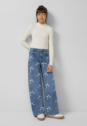 Hellbeiges Langarmoberteil kombiniert mit weit geschnittenen blauen Denim-Jeans mit Schleifenmuster. Weiße und cremefarbene Schuhe mit strukturierter Sohle.