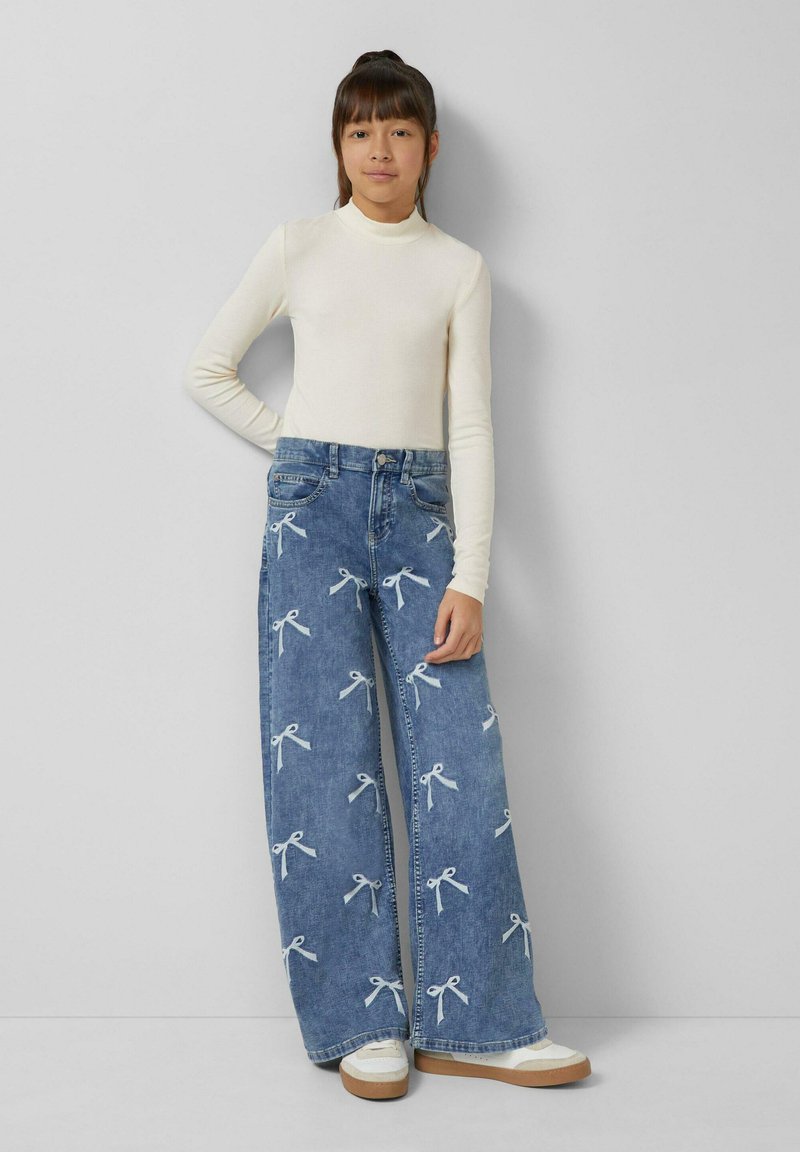 Jasnobeżowa bluzka z długim rękawem w połączeniu z szerokimi, niebieskimi jeansami z motywem kokardek. Białe i kremowe buty z teksturowaną podeszwą.