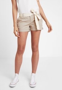 Shorts en coton beige avec une ceinture en tissu assortie, revers roulés et poches latérales. Associés à des baskets blanches montantes et un haut blanc.