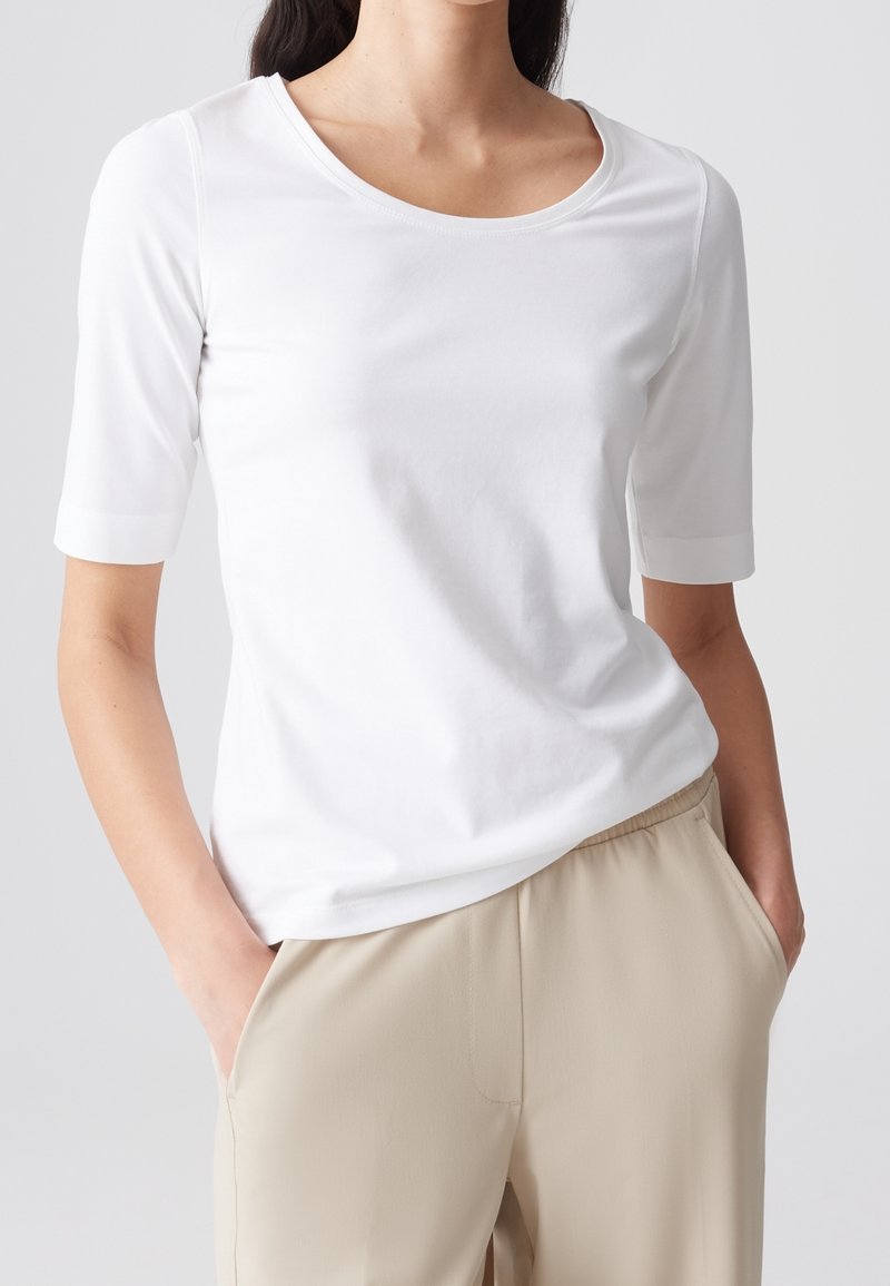 Femme portant un haut blanc uni à manches courtes et un pantalon beige, les mains dans les poches, sur un fond clair.