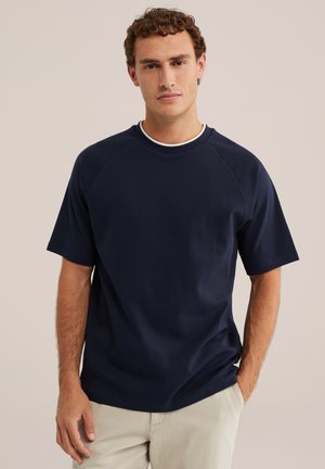 Uomo con capelli ricci che indossa una maglia blu navy a maniche corte e pantaloni beige chiaro, in piedi con una mano in tasca.