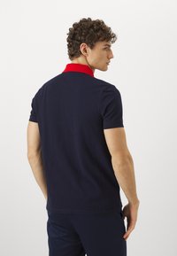 Tengeri kék pólóing piros gallérral, texturált anyagból készült, karcsú fazonú. A hátsó nézet rövid ujjakat és sima felületet mutat.