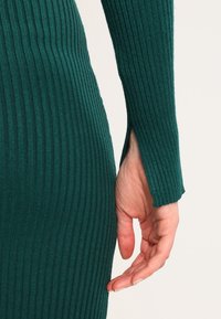 Main gauche reposant à côté du corps, portant un vêtement à manches longues en ribbed vert avec une petite fente au poignet.