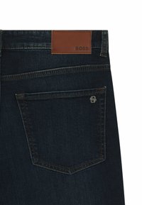 Jeans de mezclilla oscura con una etiqueta de cuero de marca, un acento de logo metálico y costuras en contraste en el bolsillo trasero.