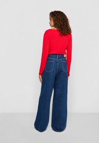 Kvinne med krøllete hår iført en rød langermet topp og høyhælte, vide blå jeans, som står mot en ensfarget bakgrunn.