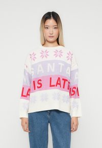 Junge Frau mit glattem Haar, die einen weißen Pullover mit pinken und roten Schneeflocken und dem Text "SANTA IST SPÄT" trägt, kombiniert mit blauen Jeans.