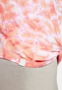 Lightweight top with a pink and orange tie-dye pattern. Features a stitched hem and a small label on the side, worn over beige leggings.